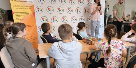 Powiększ grafikę: „Dzieci siedzą przy stołach i słuchają prowadzących podczas zajęć edukacyjnych, na stole leży gra planszowa.”