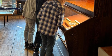 Powiększ grafikę: Uczniowie oglądają eksponaty w muzeum, stojąc przy gablocie z zabytkowymi przedmiotami