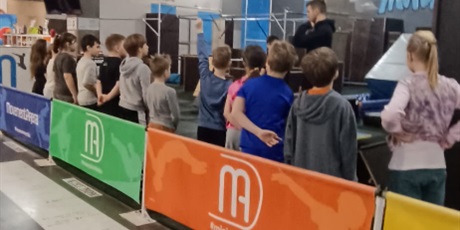 Powiększ grafikę: „Grupa uczniów słucha instruktora w hali z torem przeszkód przed rozpoczęciem zajęć ruchowych.”