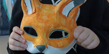 Powiększ grafikę: „Uczeń prezentuje ręcznie wykonaną maskę lisa pomalowaną na pomarańczowo z czarnymi i białymi elementami.”