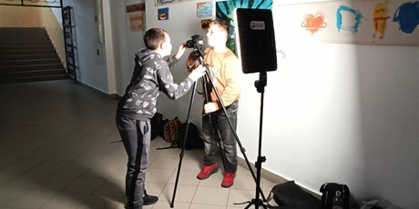 Powiększ grafikę: „Dwóch uczniów ustawia kamerę i oświetlenie w szkolnym korytarzu, przygotowując nagranie przy pracach plastycznych na ścianie.”