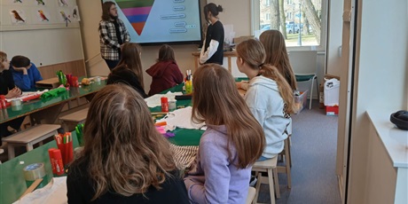 Powiększ grafikę: „Uczniowie siedzą przy stołach i słuchają prezentacji nauczycieli wyświetlanej na ekranie multimedialnym w klasie.”